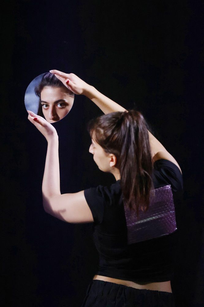 Beyond the Bubble Bühnenfoto © Ute Langkafel I Eine junge Frau steht muit dem Rücken zur Fotografin. Sie Blick in einen kleinen runden Spiegel . Ihre Augenpartie wird für die Betrachtende Person im Spiegel sichtbar, Der Hintergrund ist schwarz