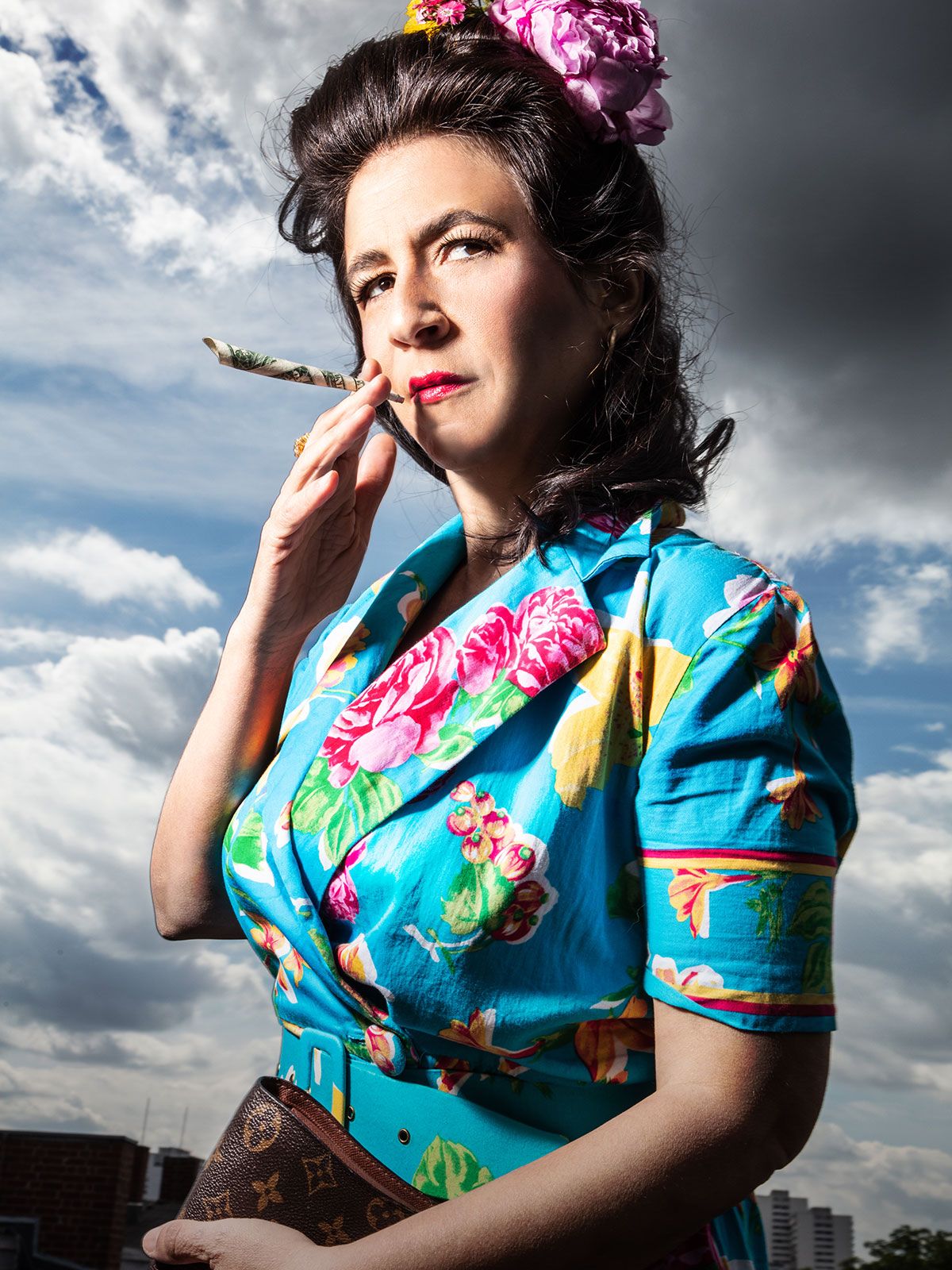 Die Schauspielerin Orit Nahmias trägt ein blaues Kleid mit Blumen. In ihre Haare gibt es auch eine Blume. Sie raucht eine Zigarette, die aus eine Geldschein gemacht ist und hält eine Louis Vuitton Tasche. The actress Orit Nahmias wears a blue dress with flowers. In her hair there is also a flower. She smokes a cigarette made of a banknote and holds a Louis Vuitton bag.
