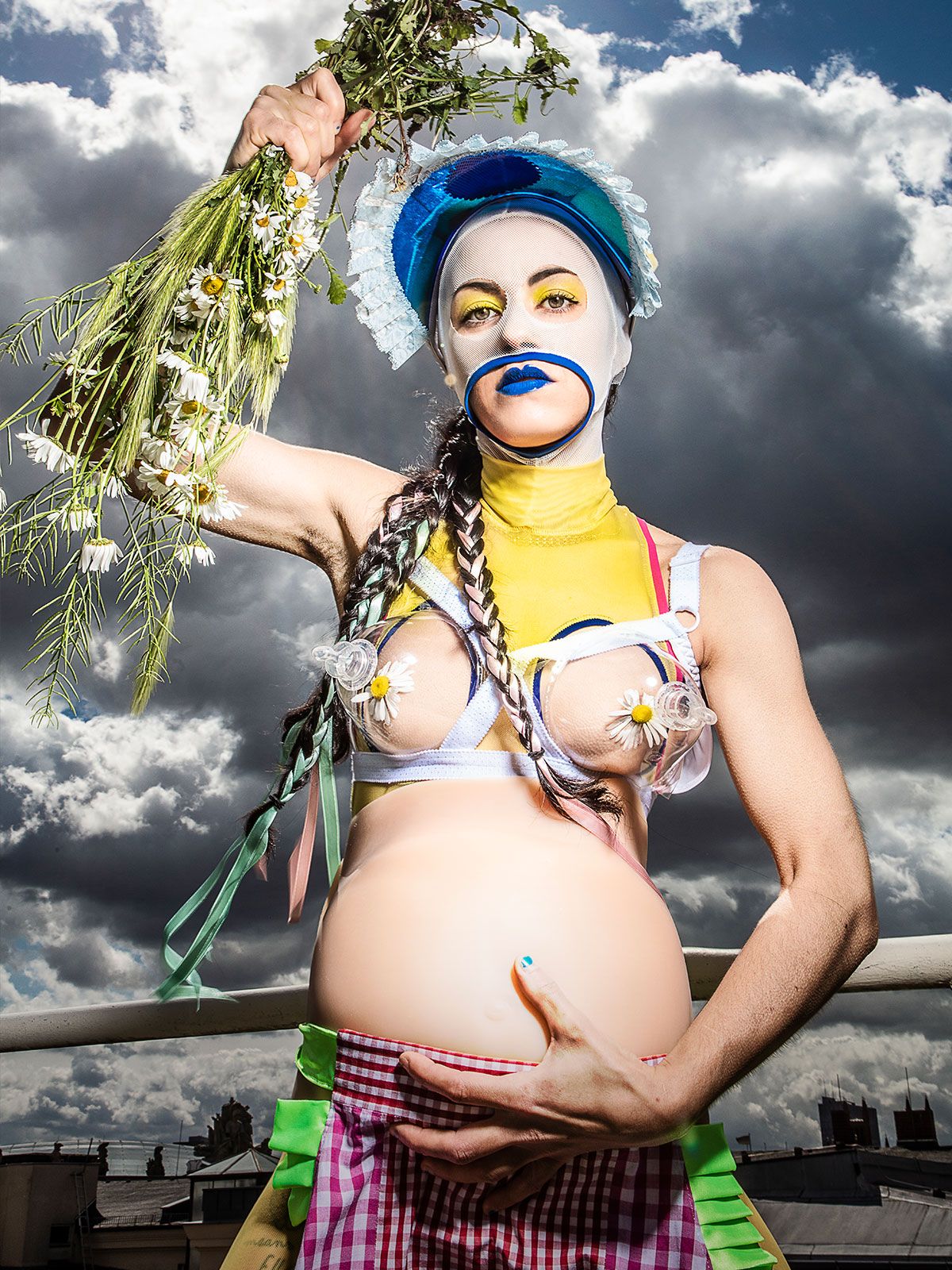 Mother Tongue I (c) Esra Rotthoff Die Schauspielerin starrt in die Kamera. Mit ihrer rechten Hand hält sie einen Strauß Gänseblümchen, mit der linken Hand hält sie ihren schwangeren Bauch. Ihre Brustwarzen sind mit Gänseblümchen und Milchpumpen bedeckt. Ihr Gesicht ist mit einer weißen Maske bedeckt, so dass nur ihre Augen und ihr Mund zu sehen sind. Ihre dunklen Haare sind geflochten und sie trägt einen blauen Hut mit weißen Rüschen.