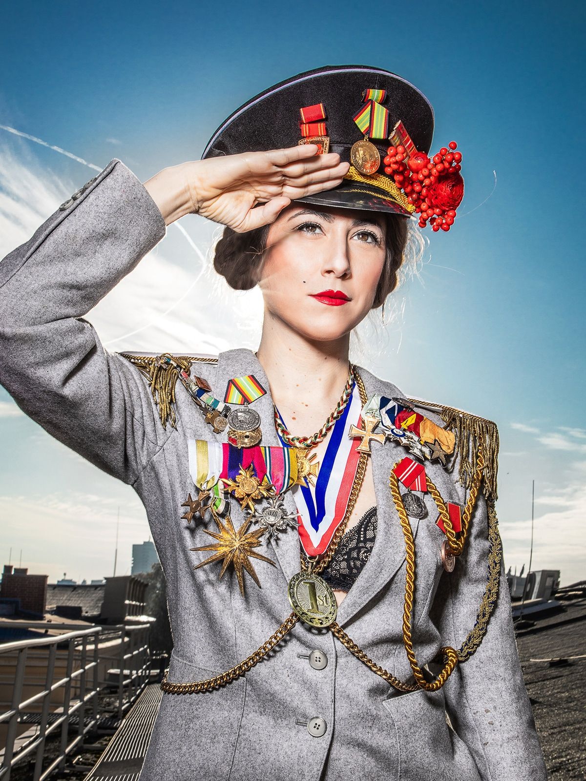SCHLACHTEN; Vidina Popov fotografiert von Esra Rotthoff; Im Zentrum des Fotos steht eine junge Frau, die ihre rechte Hand salutierend an die Stirn hält. Sie trägt eine graue Uniformjacke, an der viele Orden befestigt sind und eine Schirmmütze, an der ebenfalls Orden stecken und auch ein paar rote Blumen. Unter der Jacke ist ein schwarzer BH zu sehen und um ihren Hals hängen zwei Medaillen. Im Hintergrund sieht man einige Dächer und einen sonnigen Himmel.