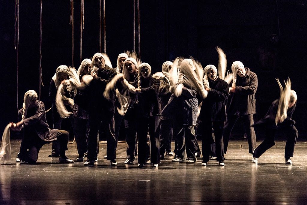 MMOJA FABRICA I Schauspieler fotografiert von Veljia Hasanbegović; 14 Schauspieler stehen auf der Bühne eng beieinander. Sie tragen alle schwarze Klamotten, weiß geschminkte Gesichter mit schwarzen Augen und Lippen sowie eine lange weiße Perücke. Die Schauspieler scheinen die Haare zu schwingen. Links sind von der Decke hängende Seile zu sehen.
