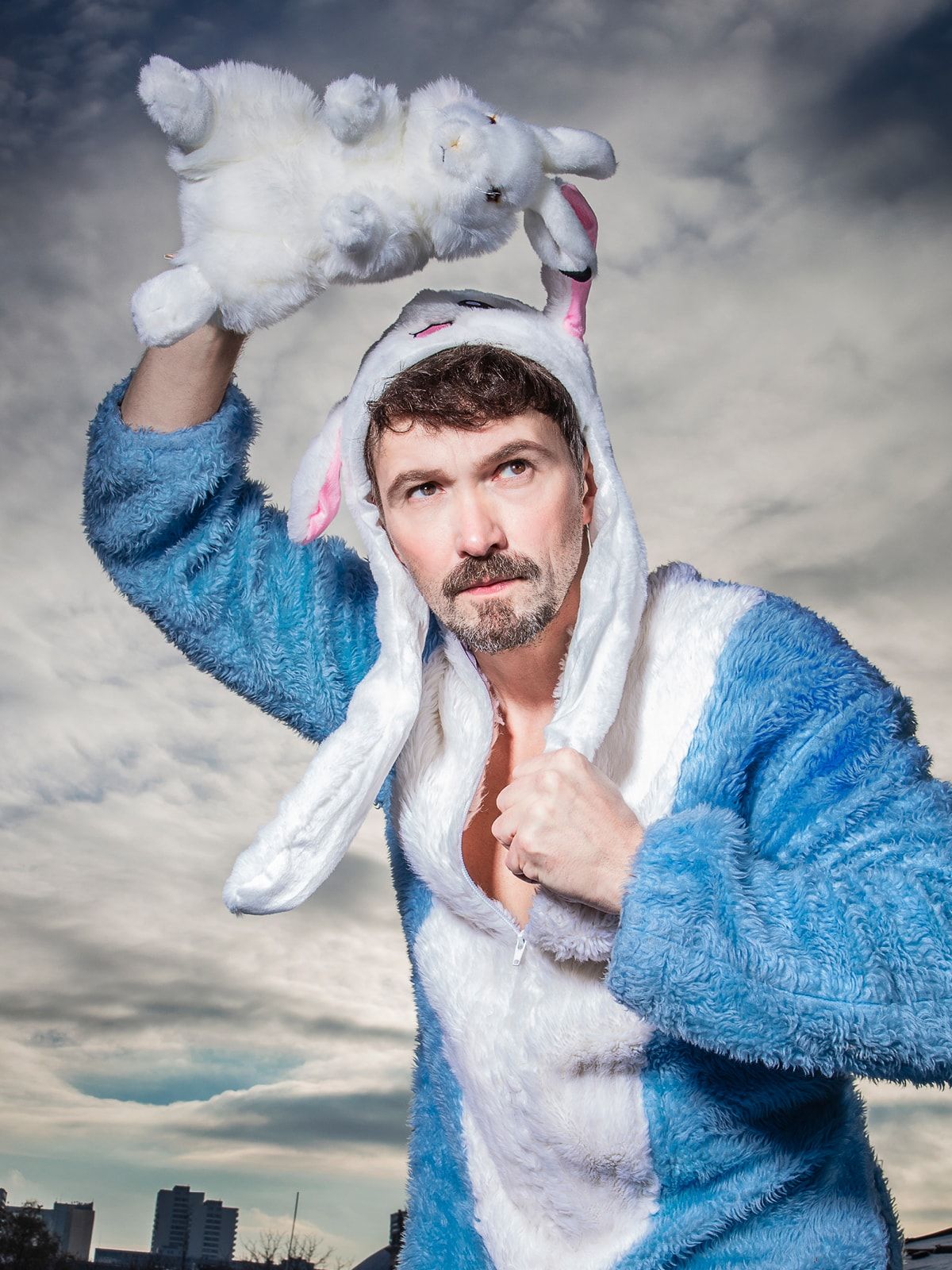 Auf dem Foto steht ein braunhaariger Schauspieler vor einem bewölkten Himmel. Er trägt einen hellblauen Plüscheinteiler, der am Bauch weiß ist. Auf dem Kopf hat er eine weiße Mütze mit rosa Ohren und langen Bändeln. Eins davon greift er mit seiner linken Hand. In seiner rechten Hand hält er ein weißes Kuscheltier-Kaninchen, das er entschlossen über seinen Kopf reckt. Sein Blick geht nach links oben.