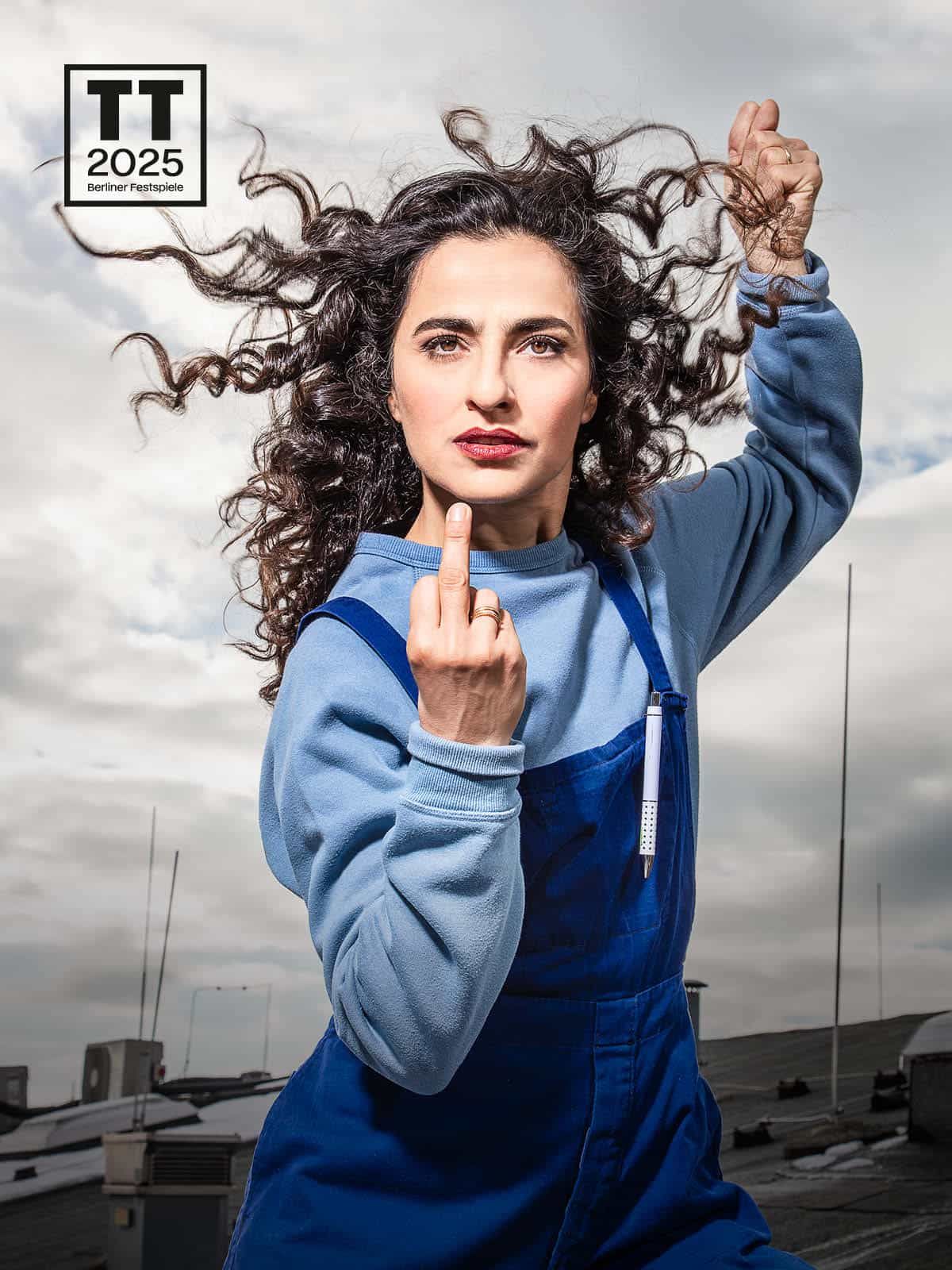 Auf dem Foto steht eine Schauspielerin auf dem Dach vor grau-bewölktem Himmel. Sie trägt einen hellblauen Pullover unter einer dunkelblauen Latzhose, in der ein Kugelschreiber steckt. Mit der rechten Hand zeigt sie den Mittelfinger frontal in die Kamera. Ihre linke Hand hält ballt sie zu einer Art Faust und hält sie hoch hinter ihren Kopf. Sie blickt konzentriert leicht an der Kamera vorbei, ihre dunkelbraunen Locken wehen ihr aus dem Gesicht.