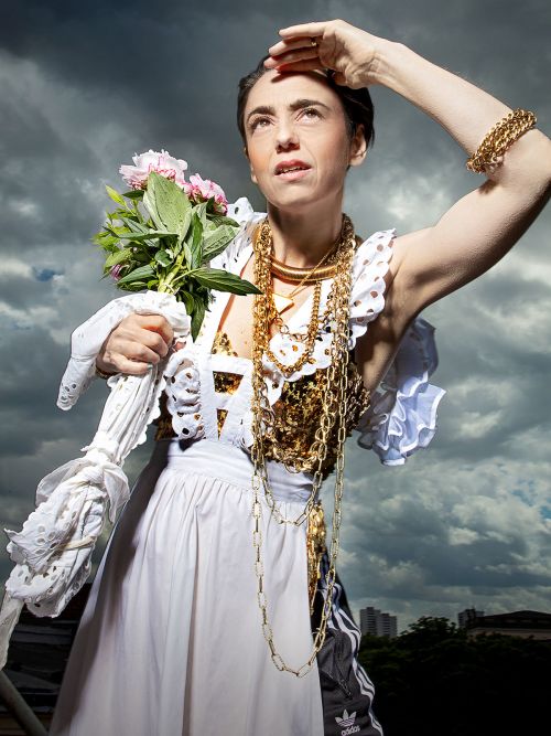 Mutter Courage und ihre Kinder (c) Esra Rotthoff Die Schauspielerin schaut nach oben. Ihr Haar ist zu einer Hochsteckfrisur gesteckt. Ihre linke Hand liegt auf ihrer Stirn. In ihrer rechten Hand befinden sich rosa Blumen und ein weißes Taschentuch. Sie trägt lange goldene Halsketten, eine weiße Schürze und darunter eine schwarze Jogginghose.
